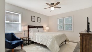 A bedroom with a bed, two bedside tables with lamps, a ceiling fan, a desk with a computer, and two windows with blinds. - CorporateHousingSavannah.com (Savannah)