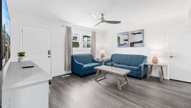 Una habitación moderna con techo blanco, ventilador de techo, dos sofás azules, una mesa de centro de madera y una mesita auxiliar con lámpara.
