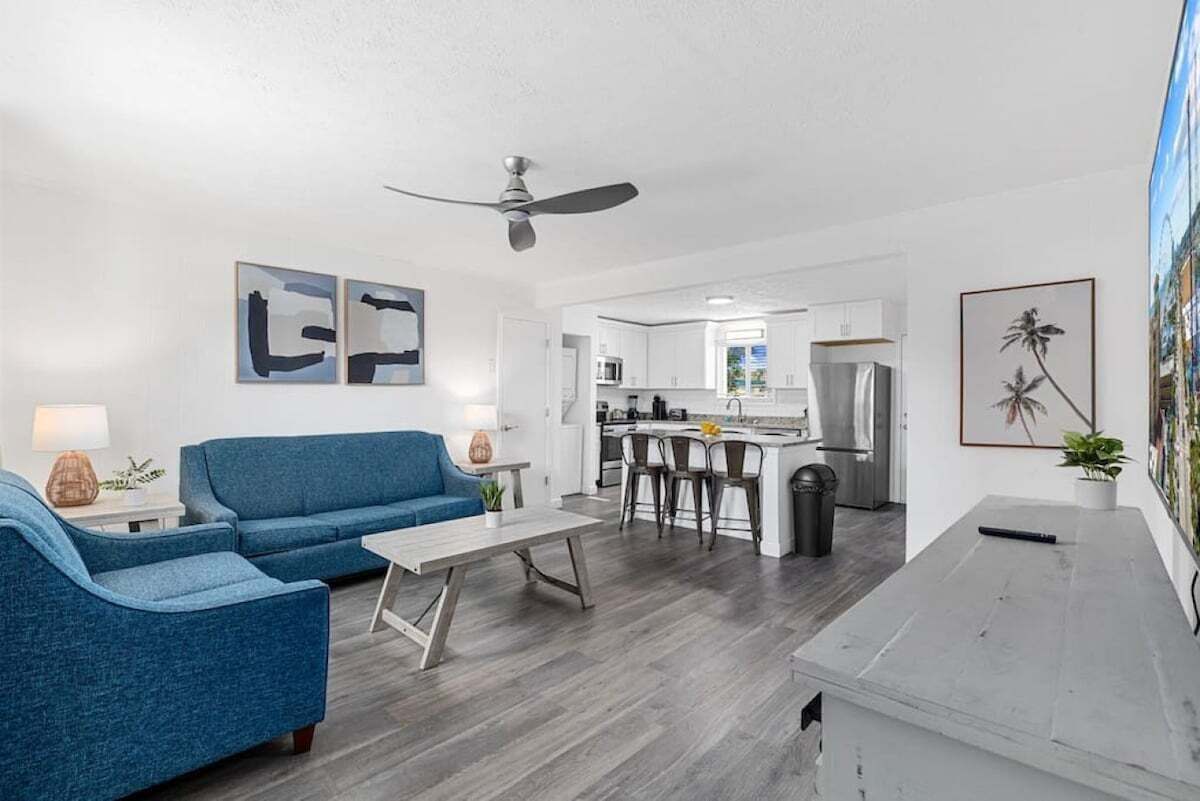 Een moderne woonkamer met een blauwe bank, een salontafel en een plafondventilator. Er is een keuken met witte kasten en een barkruk.
