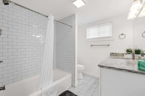 A modern bathroom with white subway tiles, a shower with a curtain, a toilet, a sink with a granite countertop, and a window with blinds.