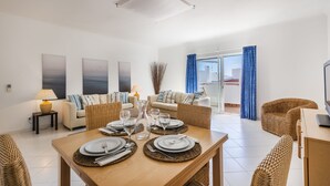 A modern living room with a dining area set for a meal, featuring a sofa, a coffee table, and a TV on a wooden cabinet. - Prainha Family Holidays (Portimão)