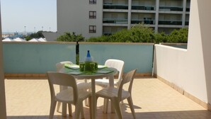 Ein Balkon mit rundem Tisch und vier Stühlen, einer Wasserflasche und einer grünen Matte. Der Balkon hat einen gefliesten Boden und Blick auf ein mehrgeschossiges Gebäude mit Balkonen.