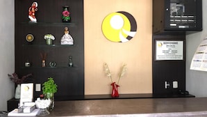 Un mostrador de recepción de hotel con una zona de registro, un panel informativo en la pared y diversos objetos decorativos, incluyendo un jarrón con flores y una planta pequeña.