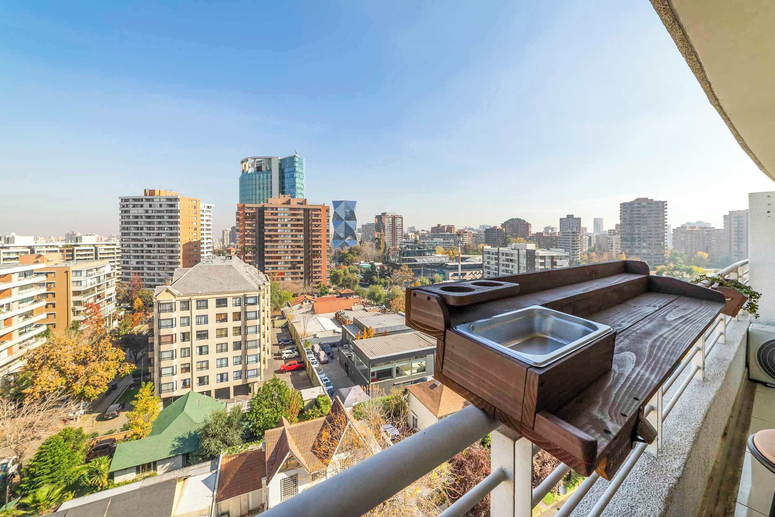Un balcón con banco de madera y lavamanos, con vista a un paisaje urbano de varios edificios.