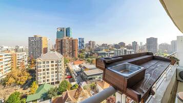 Un balcón con banco de madera y lavamanos, con vista a un paisaje urbano de varios edificios.