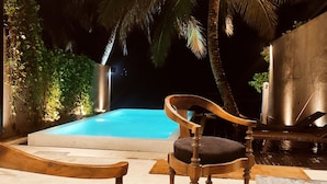 A pool area with a wooden chair and a small table, surrounded by greenery and palm trees. - Villa Reef Talpe (Unawatuna)