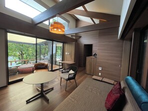 Un intérieur moderne avec une table ronde, deux chaises, un canapé et un téléviseur. De grandes fenêtres offrent une vue sur la verdure environnante.