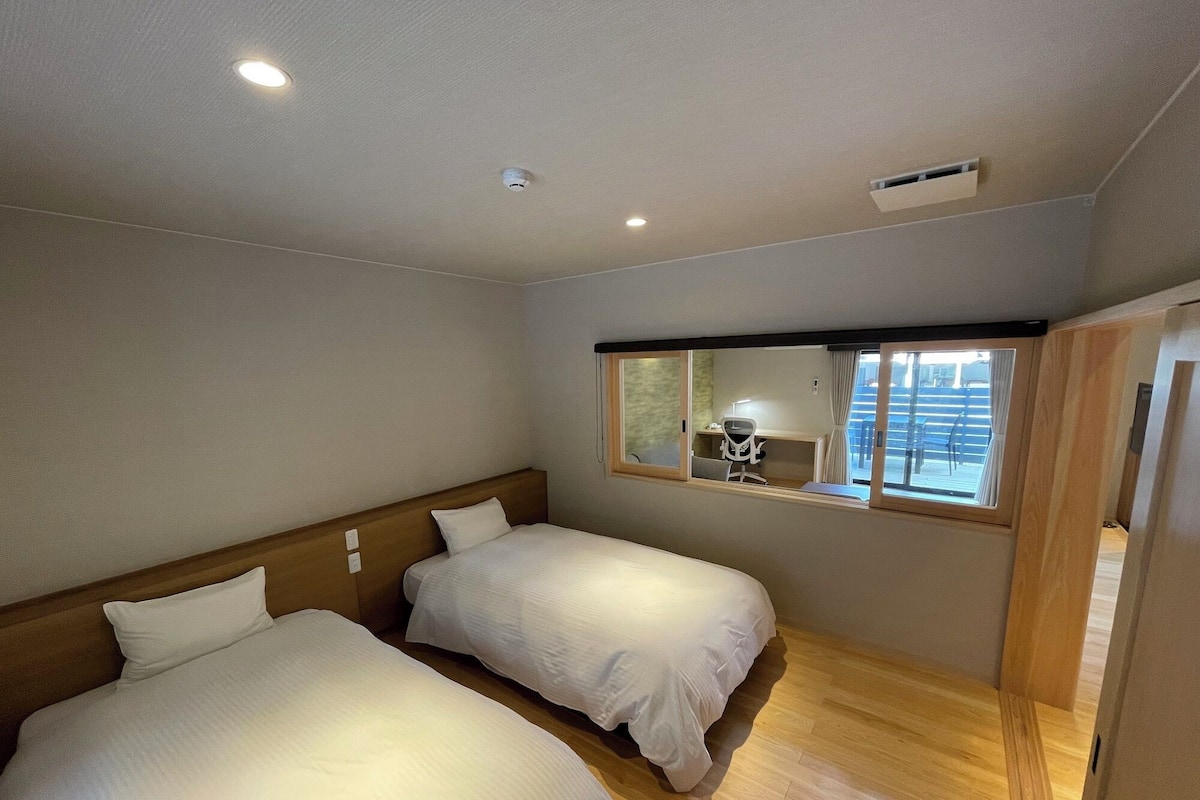 Ein Hotelzimmer mit zwei Betten, einem hölzernen Kopfteil, einem Schreibtisch mit Stuhl, einem Fenster mit Vorhängen und einer Deckenleuchte.