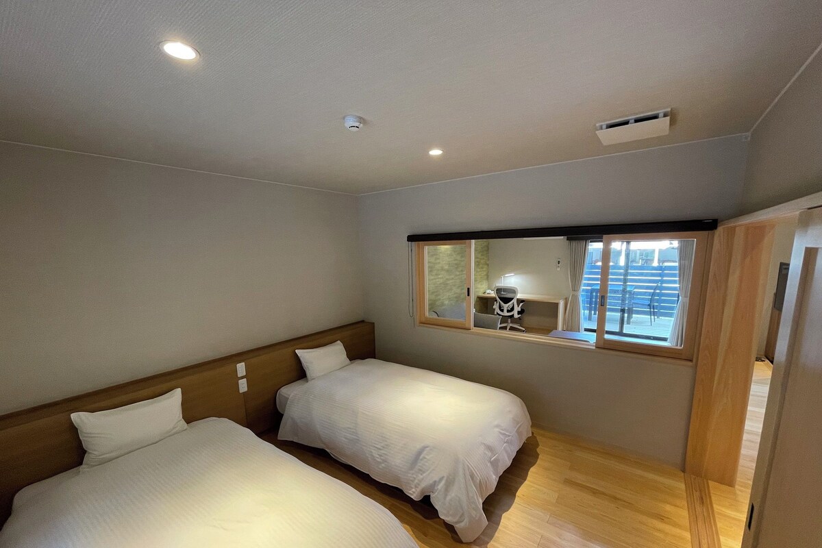 A hotel room with two beds, a wooden headboard, a desk with a chair, a window with curtains, and a ceiling light.