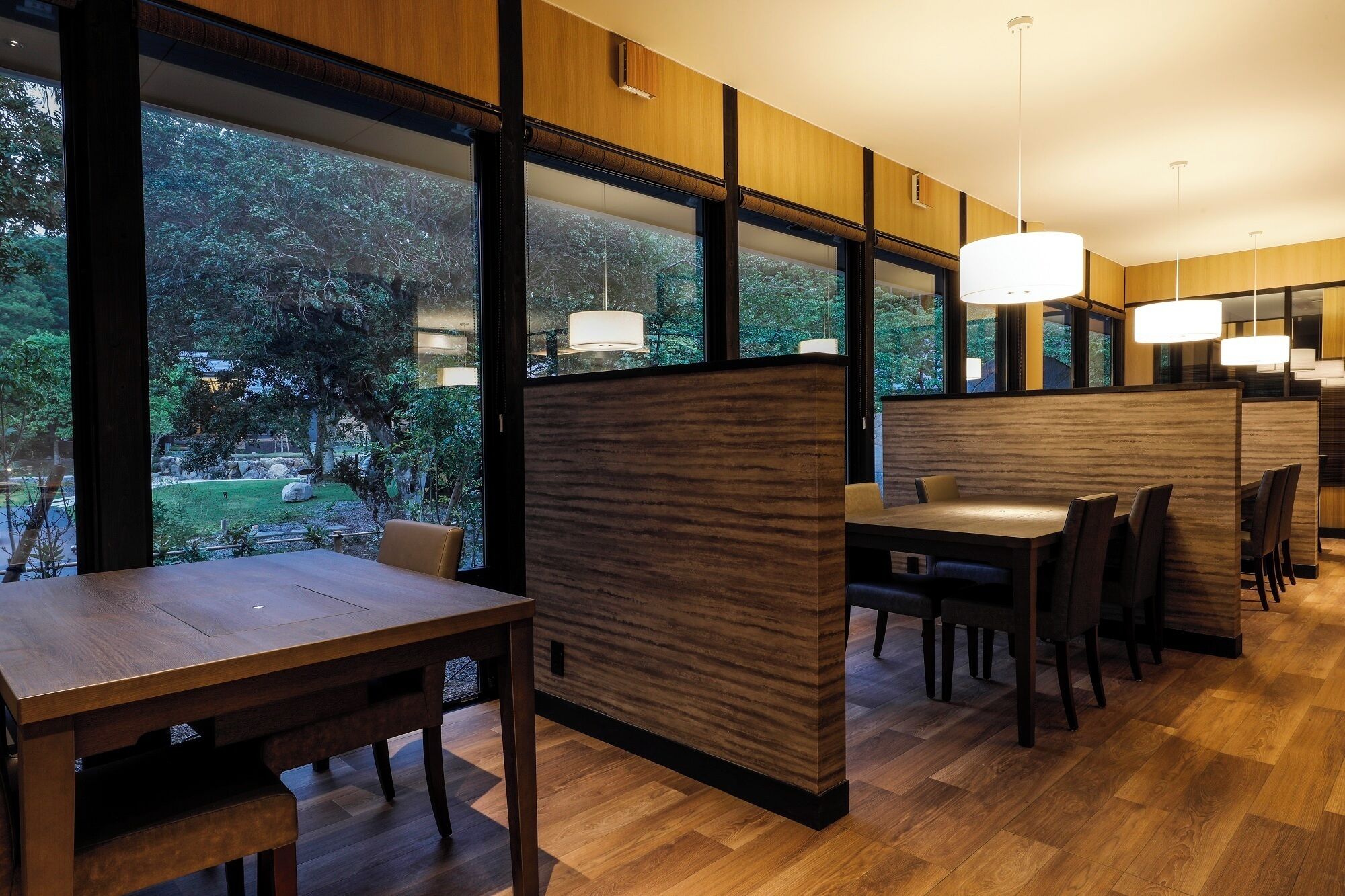 A modern dining area with wooden panel partitions, large windows offering a view of a garden, and pendant lighting.