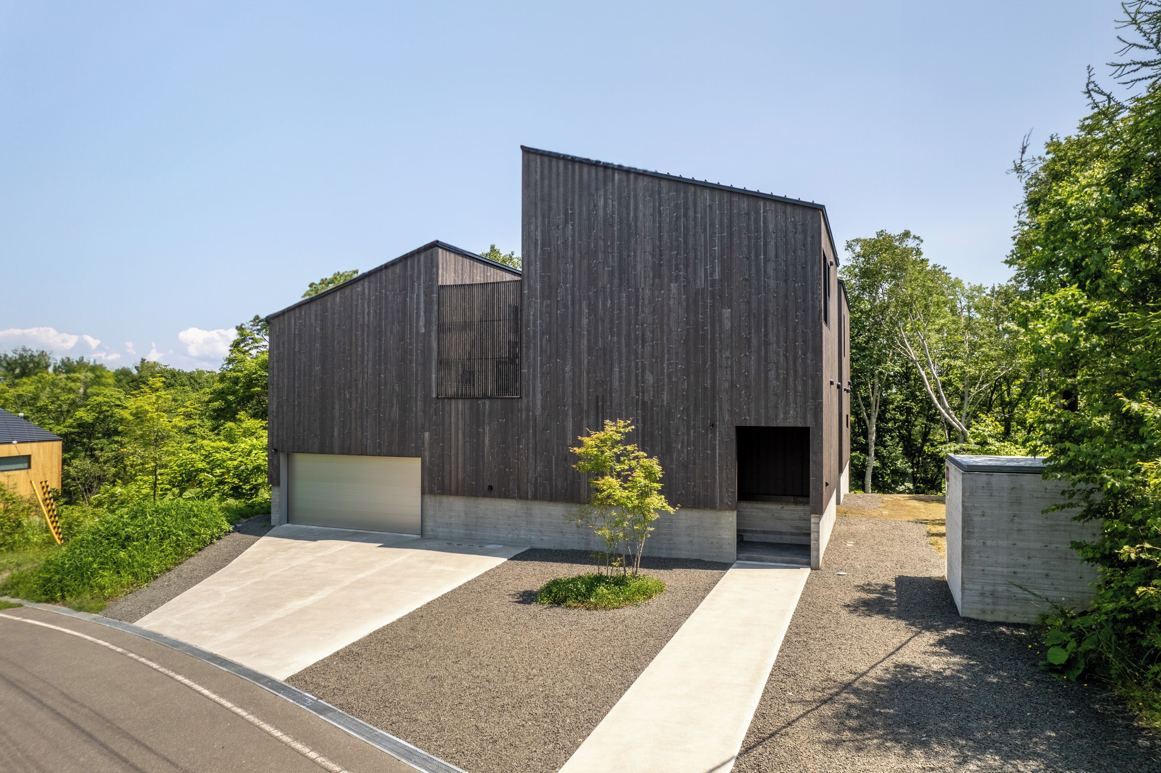 A modern building with a sloped roof, a small tree in the center, and a gravel driveway.