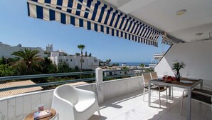 Ein Balkon mit weißen Stühlen, einem Tisch mit einer Vase und Weingläsern sowie Blick auf das Meer und Gebäude.