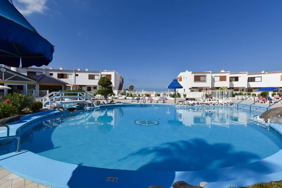 A swimming pool with a 1.40 m depth marker, surrounded by white buildings with balconies and blue umbrellas.