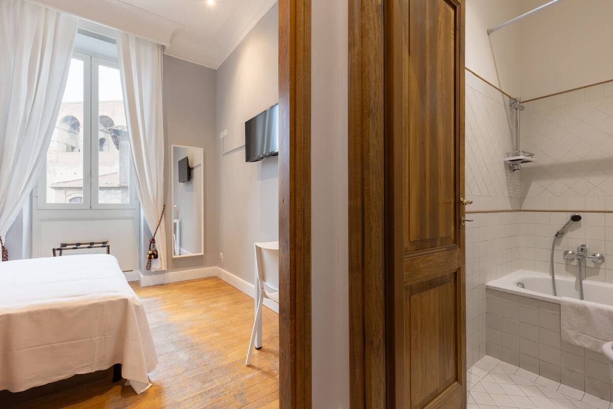 A hotel room with a wooden floor, a bed, a TV mounted on the wall, and a shower area.