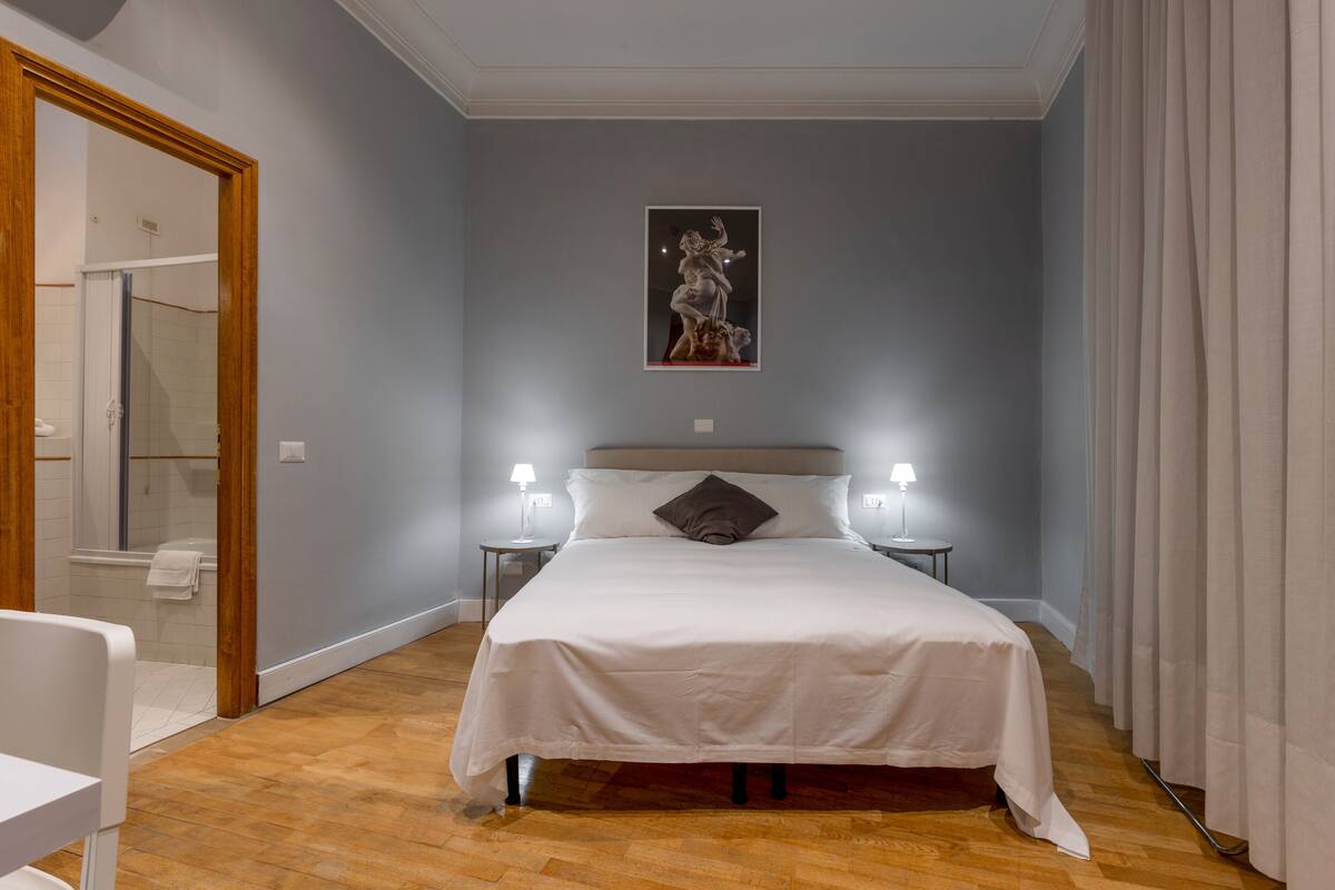 A bedroom with a bed, two bedside lamps, a nightstand, a wooden floor, a door, and a wall-mounted artwork.