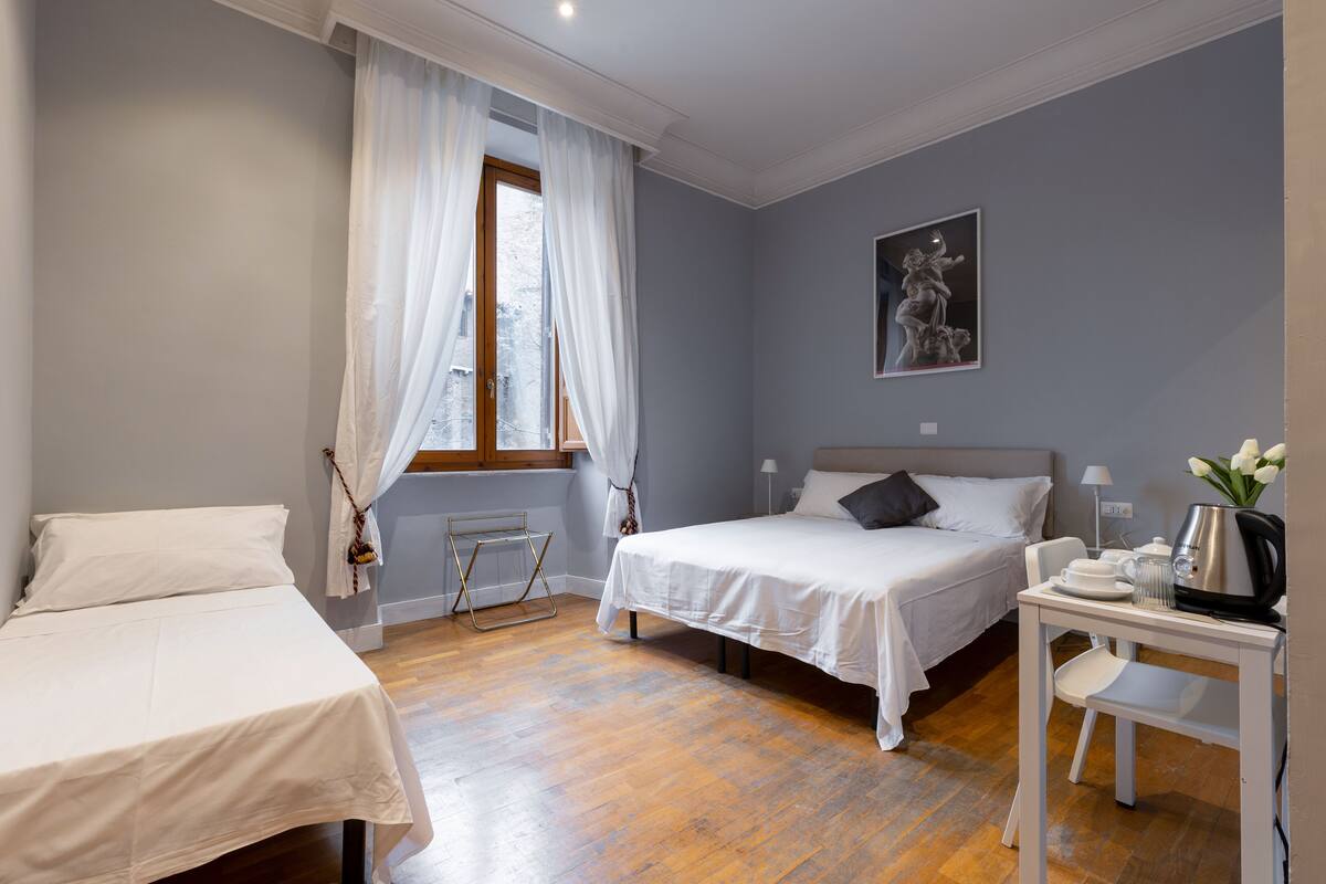 A hotel room with two beds, a small table with a kettle, and a window with curtains.