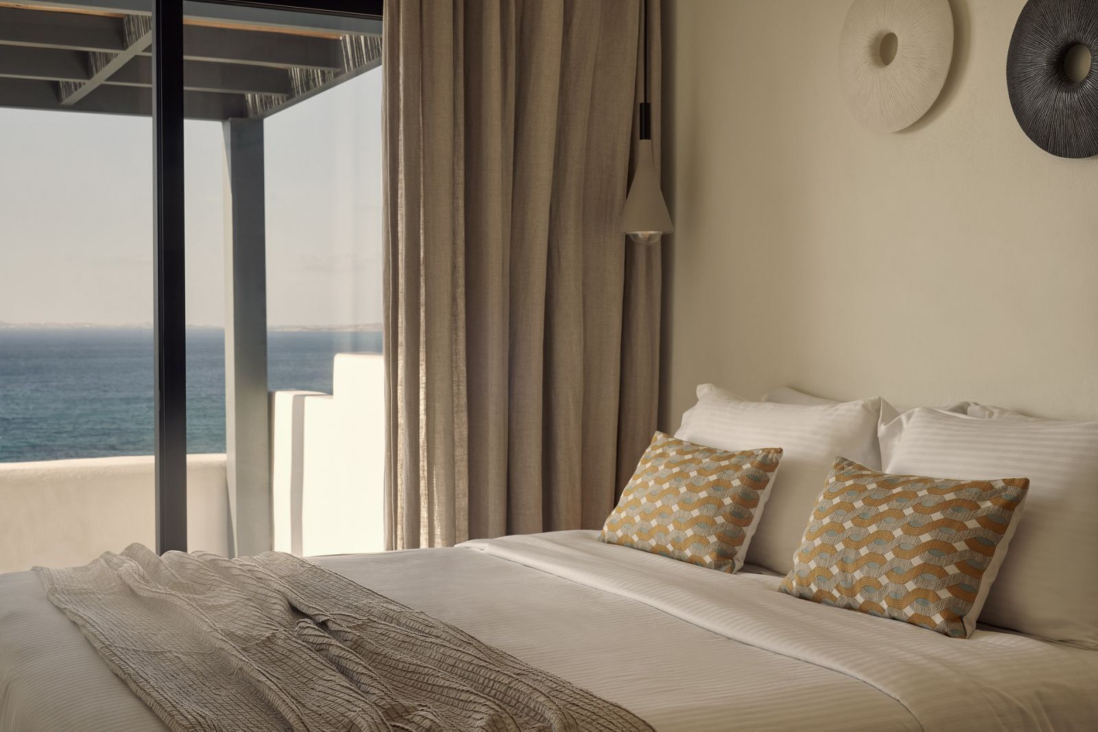 A bedroom with a bed, two patterned pillows, a throw blanket, a hanging light, and a view of the ocean.