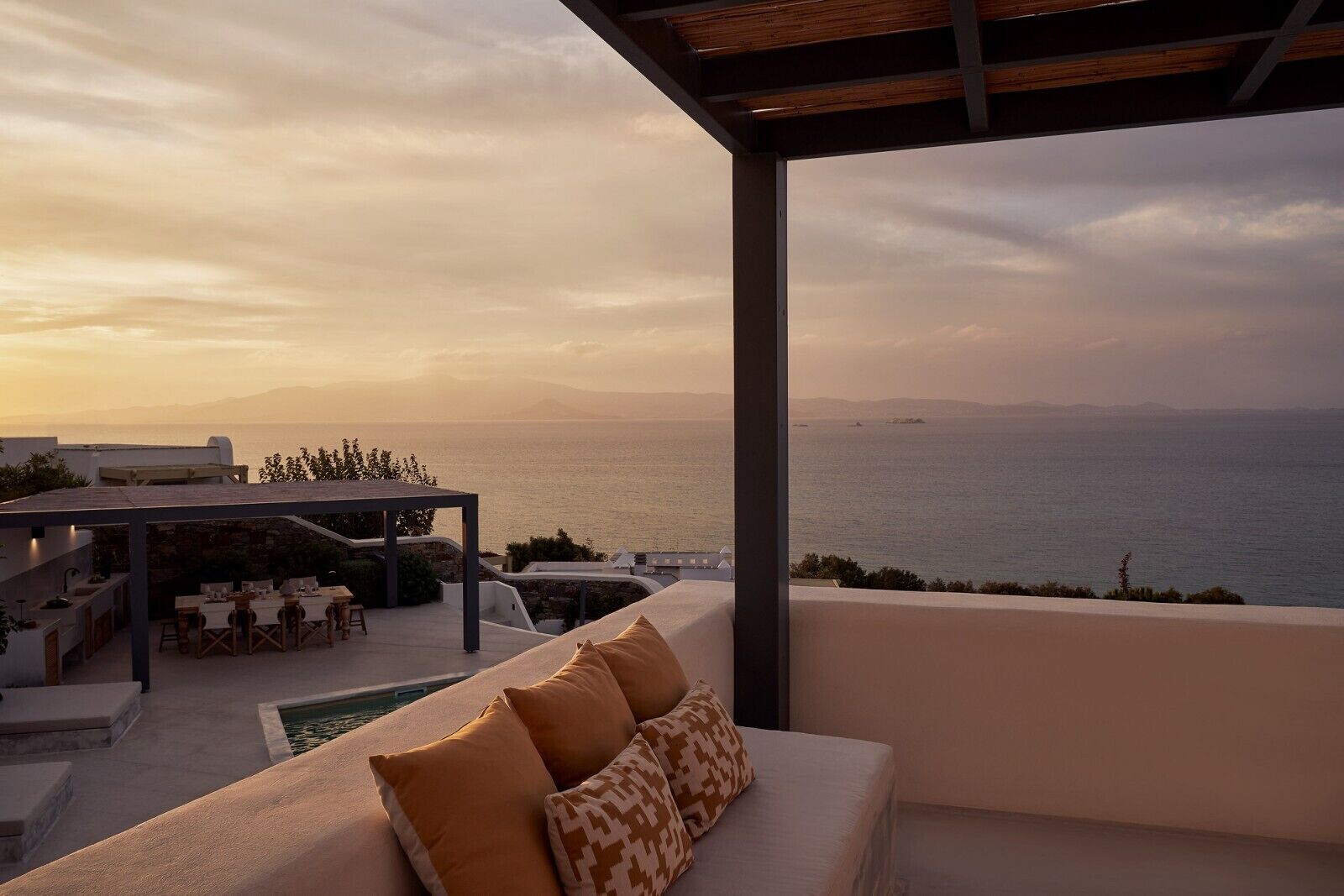 A modern outdoor seating area with a pool, overlooking a scenic ocean view at sunset.