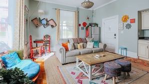 A cozy living room with a sofa, coffee table, and ottomans. There is a dining table with chairs, a small kitchen area, and a window with curtains. - The Bohemian Suite (Staunton)