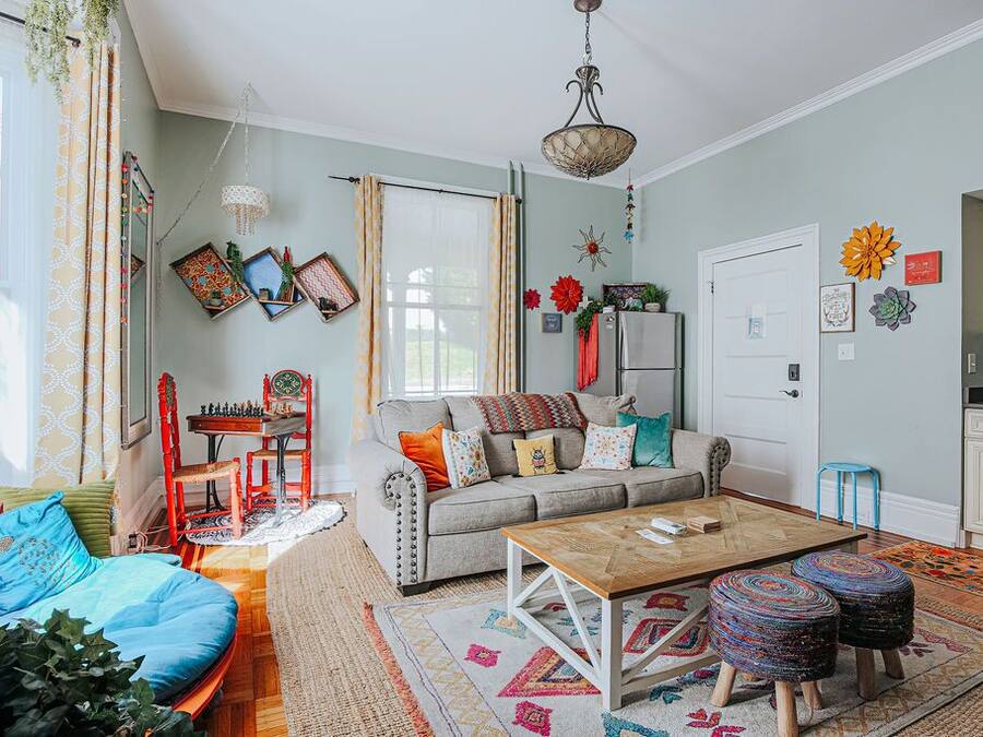 A cozy living room with a sofa, coffee table, and ottomans. There is a dining table with chairs, a small kitchen area, and a window with curtains.