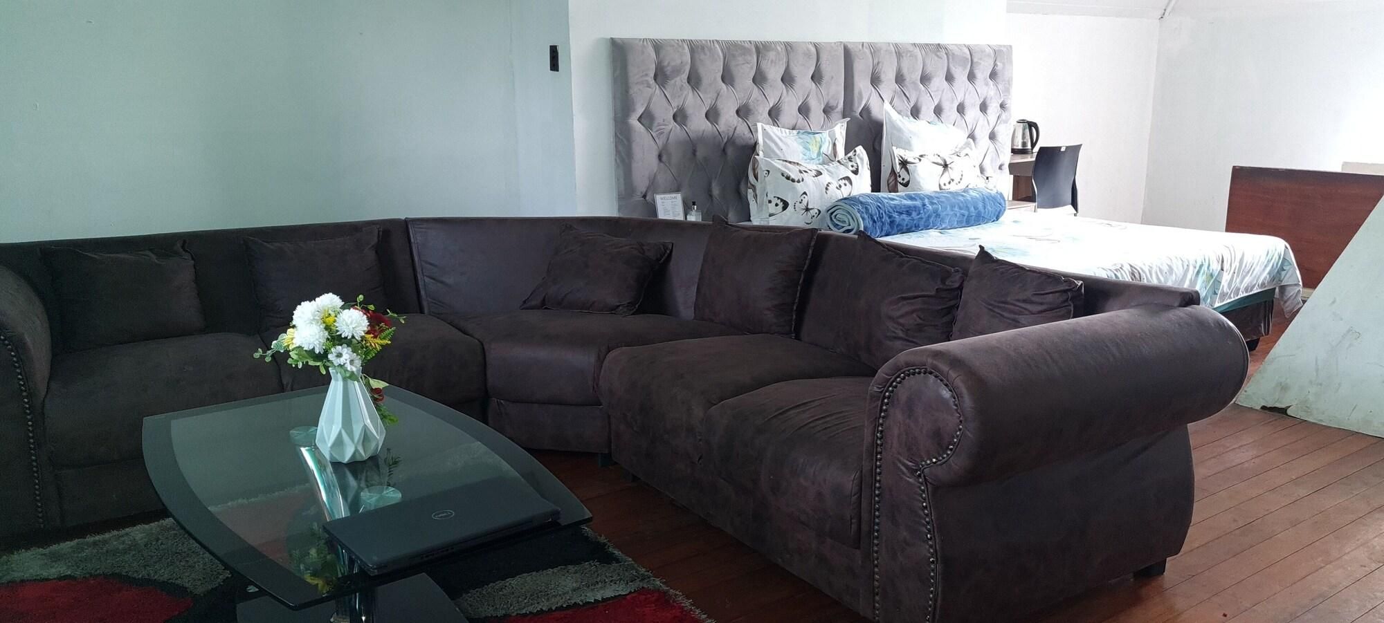 Una sala de estar con un sofá seccional, una mesa de centro de vidrio con un florero y una cama con cabecera capitonada.