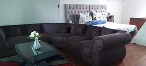 Un salon avec un canapé d’angle, une table basse en verre ornée d’un vase de fleurs et un lit doté d’une tête de lit capitonnée.