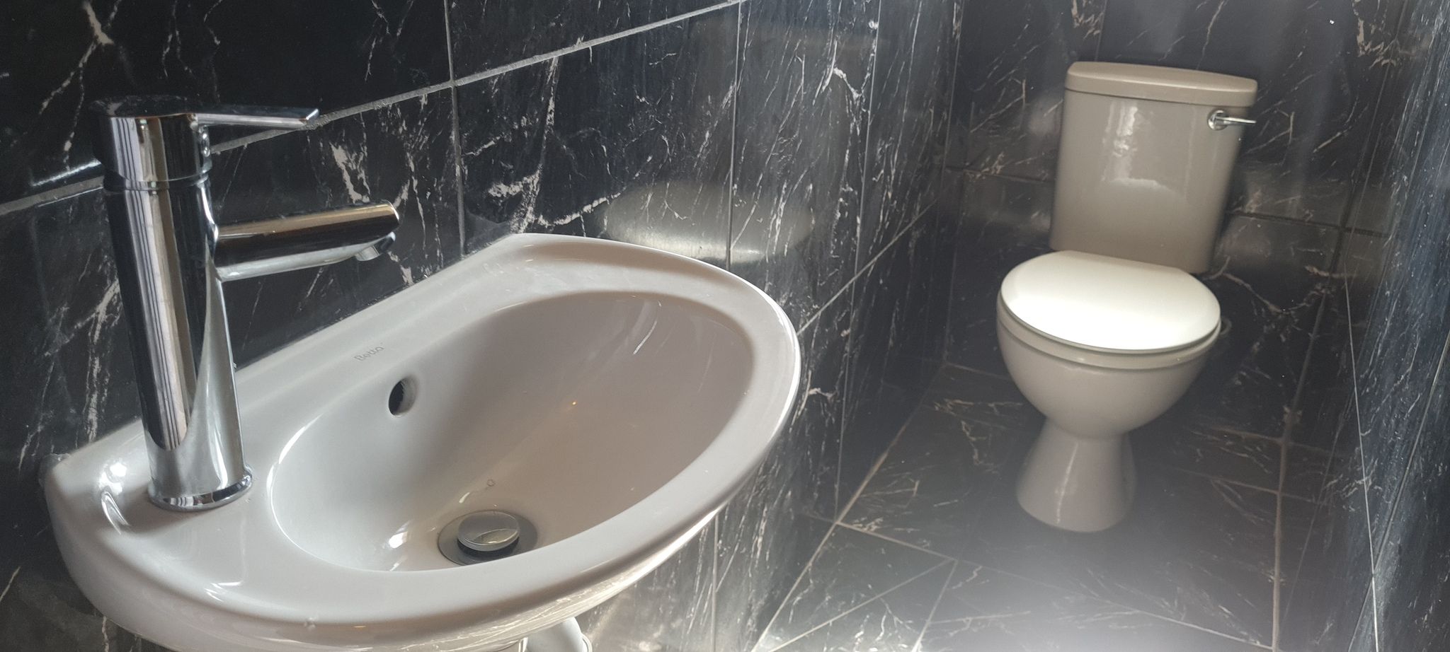 Un baño con lavamanos y retrete blancos, paredes de mármol y grifo cromado.