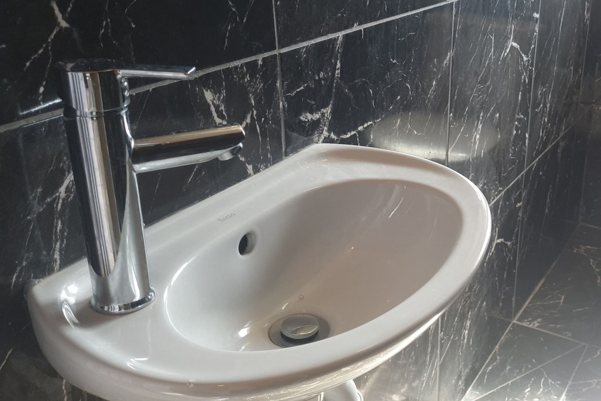 Un baño con lavamanos y retrete blancos, paredes de mármol y grifo cromado.