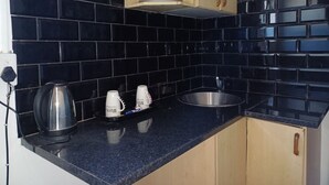 Una cocina con baldosas negras, un lavamanos de acero inoxidable, una tetera y dos tazas sobre la encimera.
