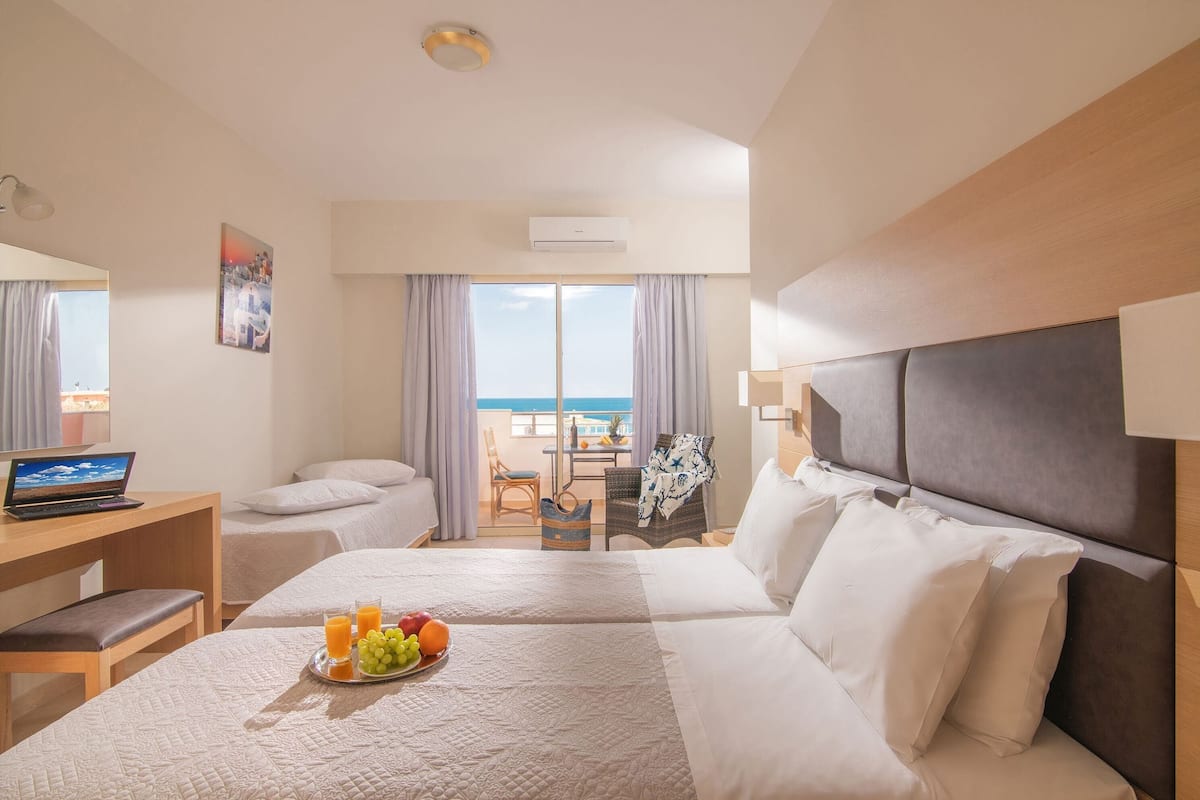 Ein Hotelzimmer mit Bett, Schreibtisch mit Laptop, Frühstückswagen mit Obst und Meerblick durch große Fenster.