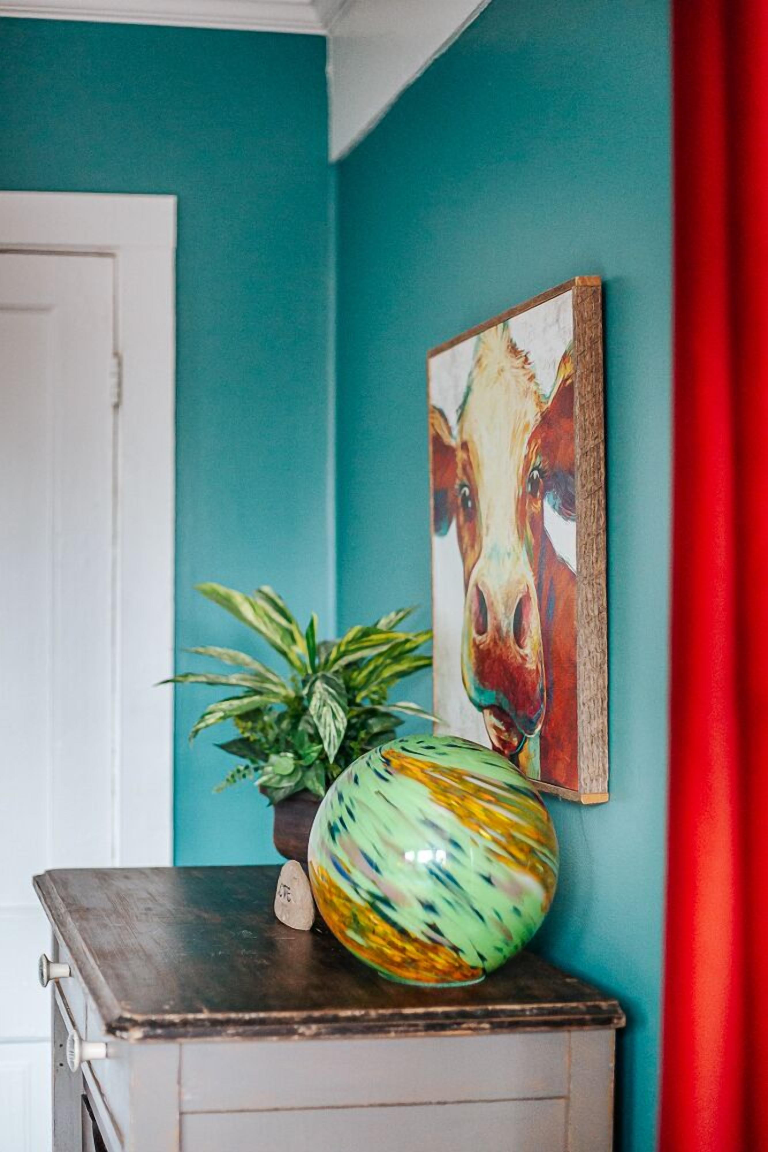A teal wall, a wooden cabinet, a potted plant, a colorful vase, and a framed picture of a cow.