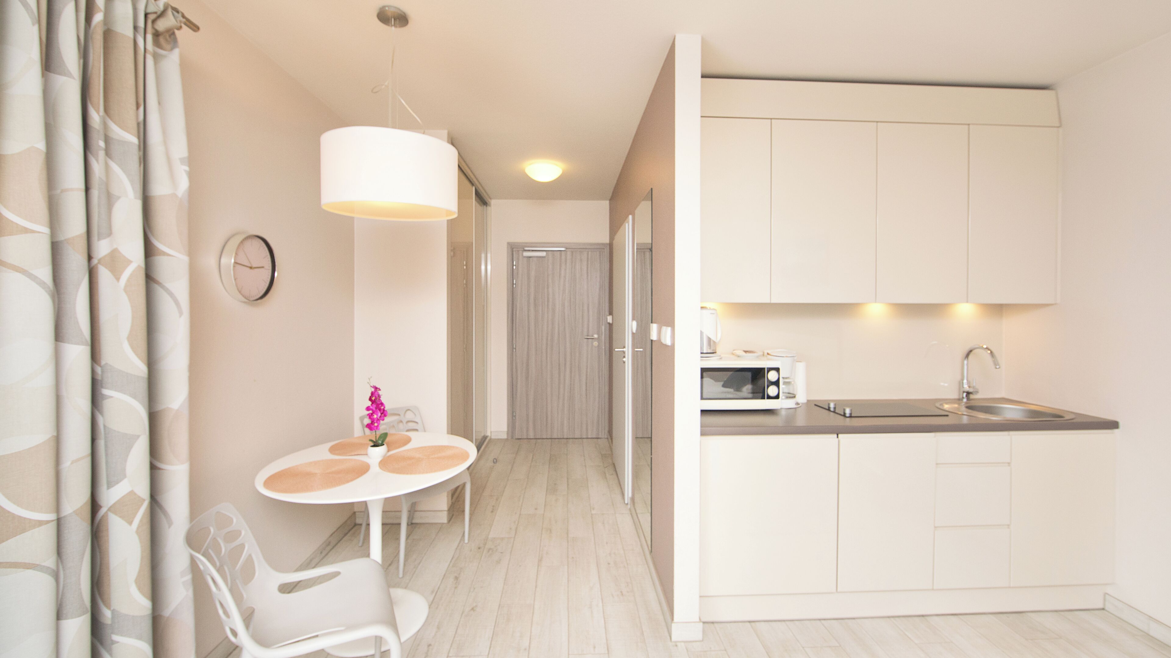 A modern kitchen with white cabinets, a microwave, and a sink. There is a dining area with a table and chairs, and a clock on the wall.