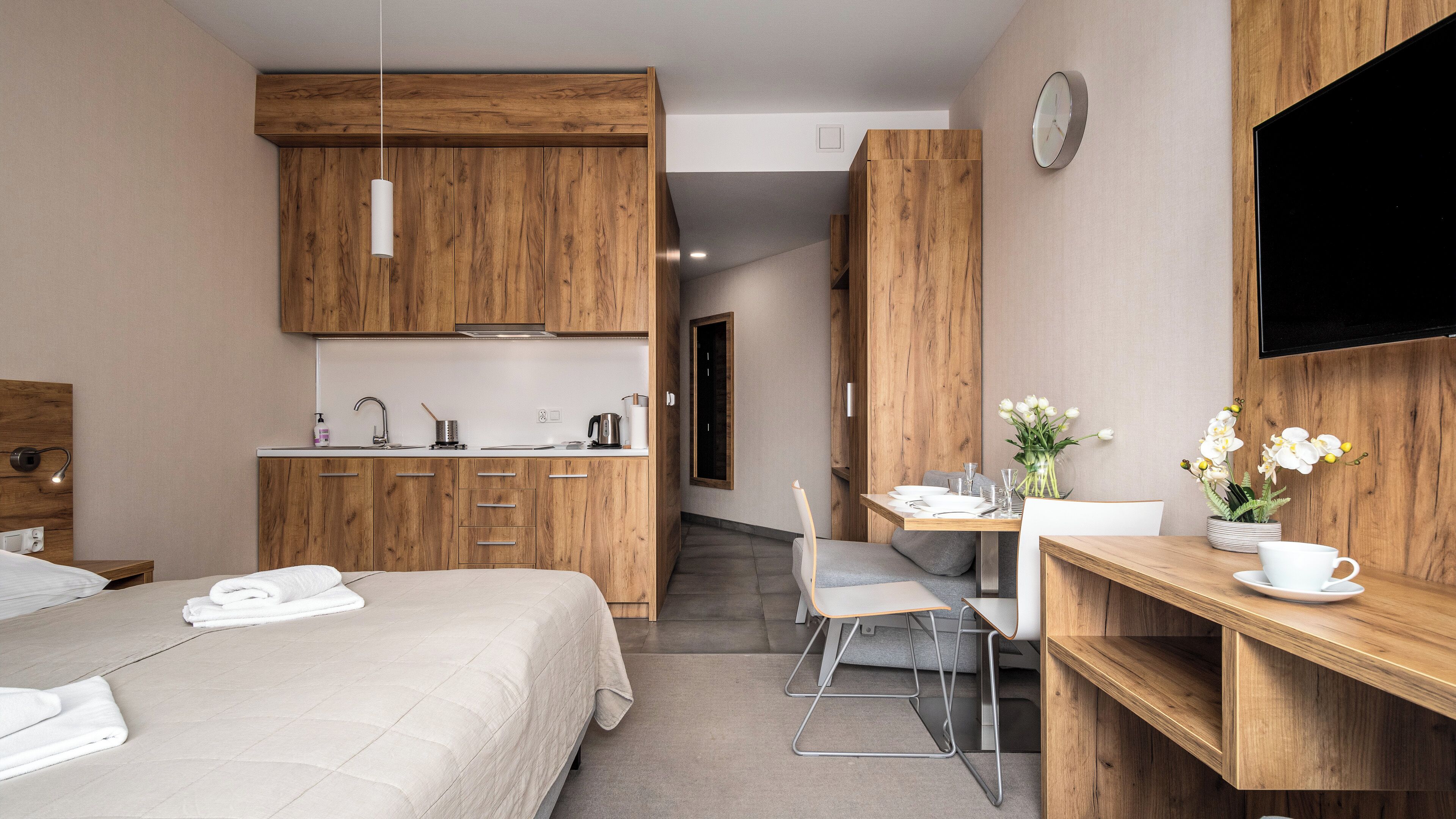 A compact hotel room with a bed, a kitchenette, a dining area, and a TV mounted on the wall.