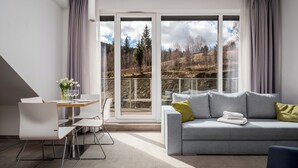 A modern living room with a grey sofa, a dining table with chairs, and a view of a forest through large windows. - Aparthotel Panorama – Czarna Góra Resort | Sun & Snow (Sienna)