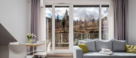 Una sala de estar moderna con un sofá gris, una mesa de comedor con sillas y vistas a un bosque a través de amplios ventanales.