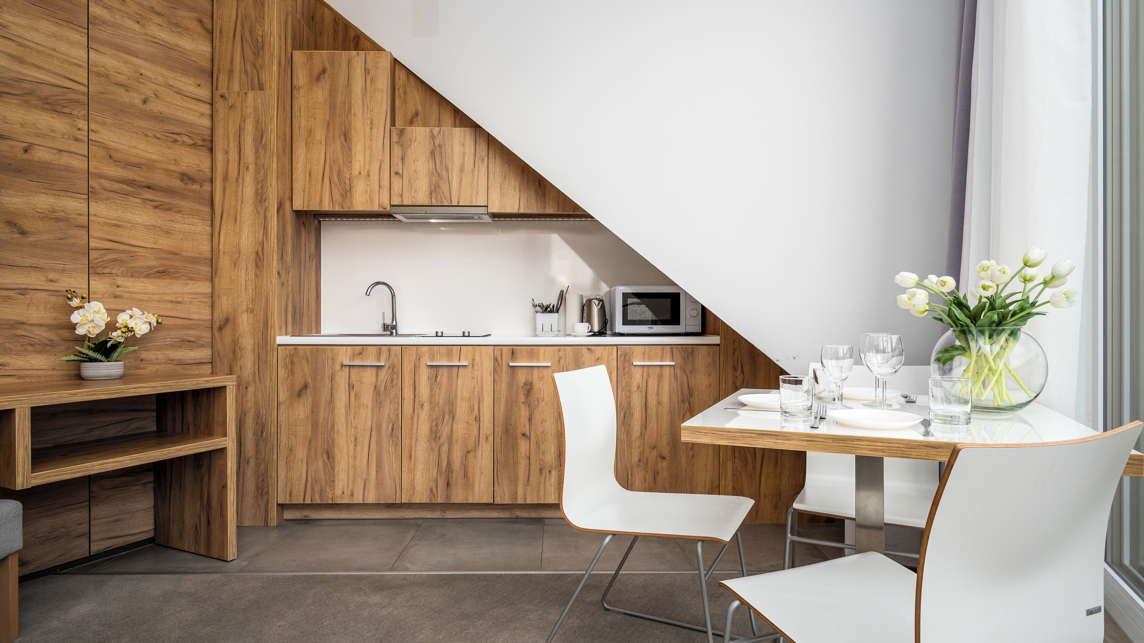 A modern kitchen with wooden cabinetry, a white countertop, and a dining area with white chairs and a table set for two.