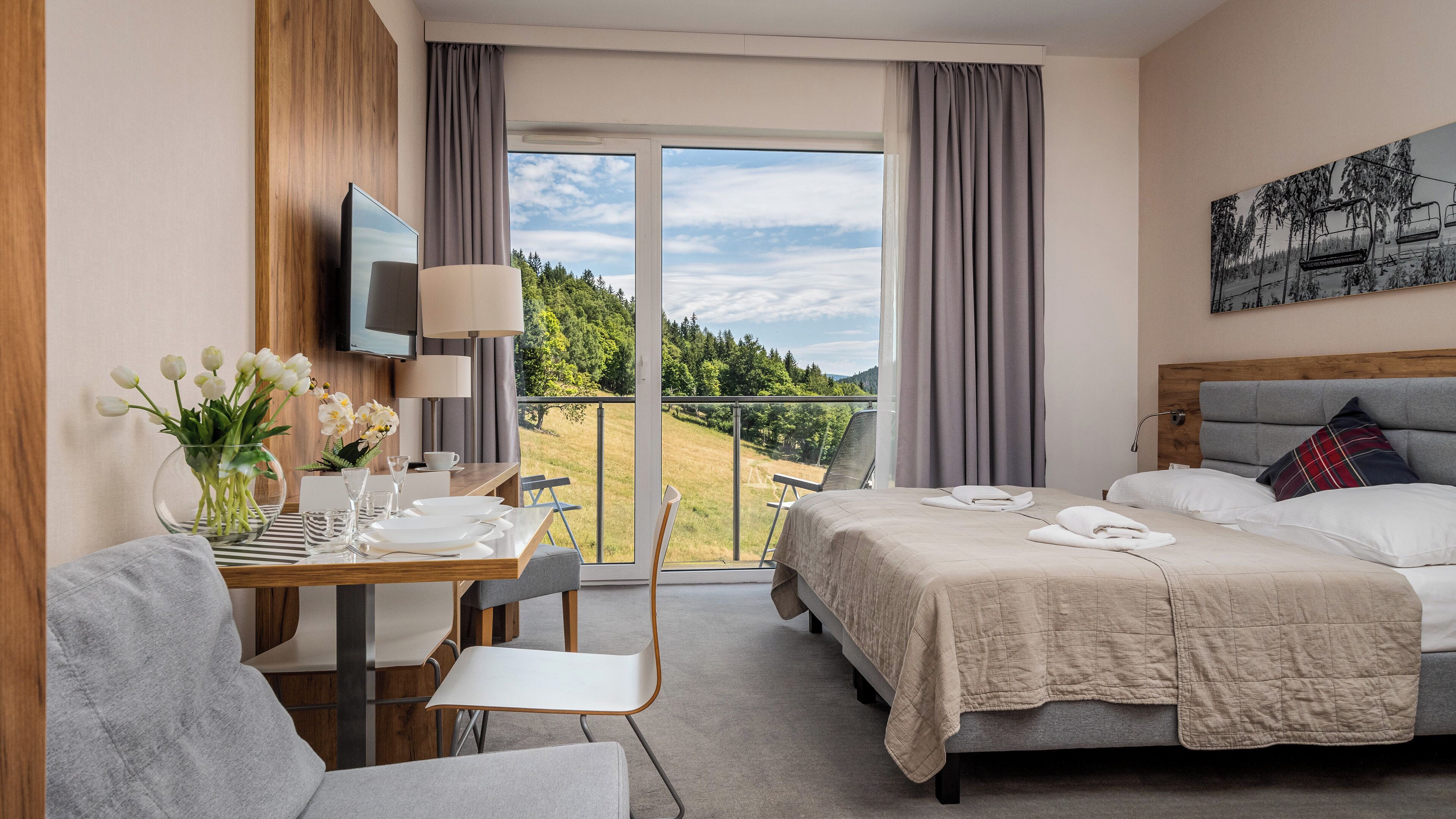 Une chambre d’hôtel comprenant un lit, un bureau avec une chaise, une télévision et un balcon offrant une vue sur les arbres et les collines.