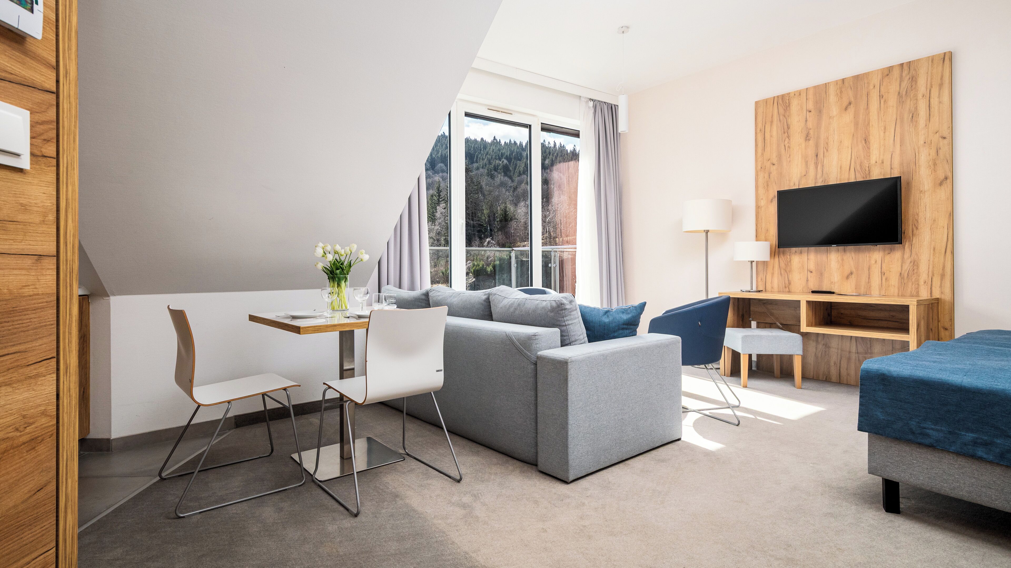 A modern hotel room with a large window, a flat-screen TV, a sofa, a desk, and a chair.