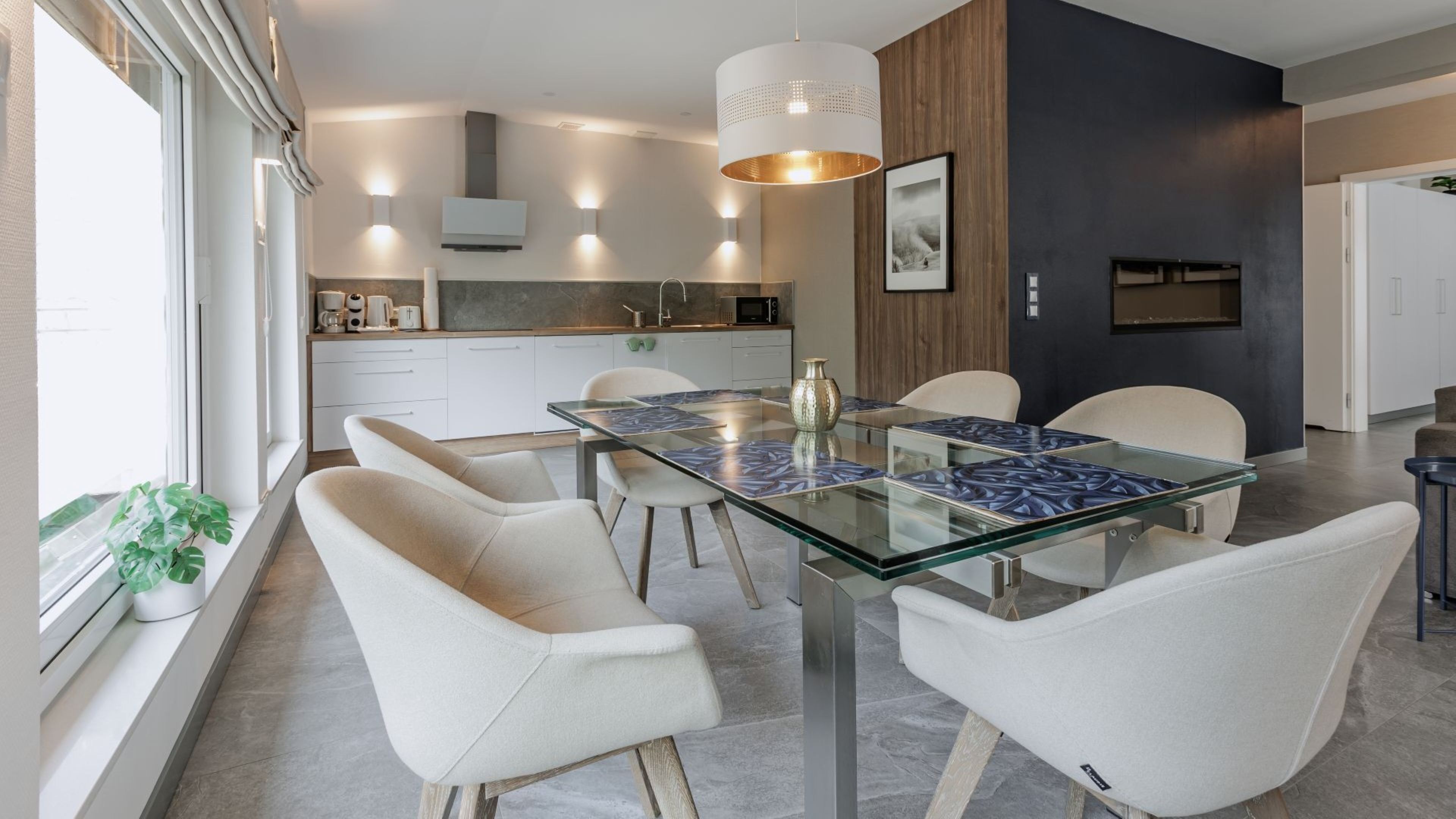 Un comedor moderno con una mesa de vidrio y sillas blancas, una cocina al fondo y un ventanal con una planta.