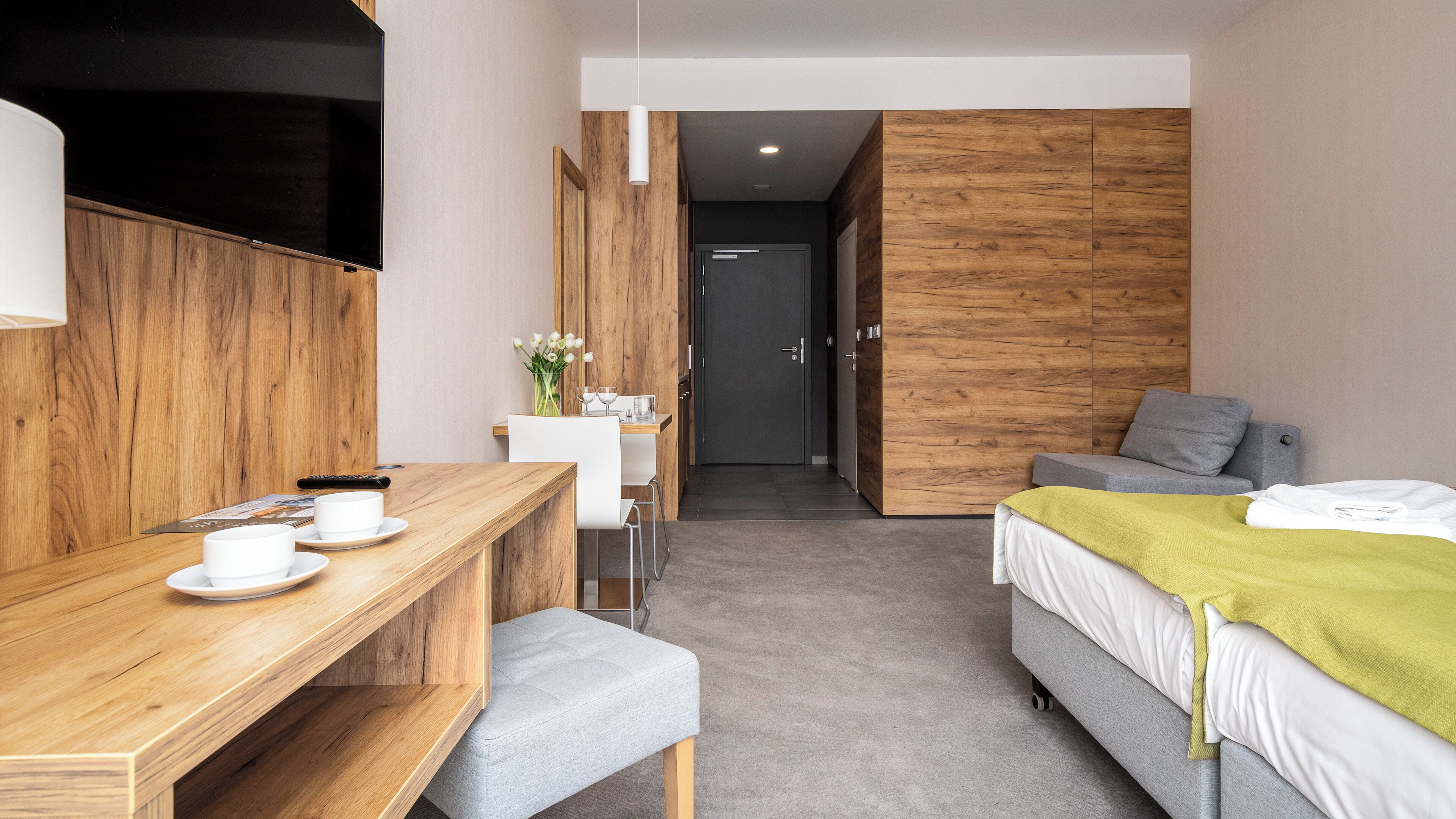 A modern hotel room with a wooden desk, a bed with a green blanket, a grey sofa, and a small table with a vase of flowers.
