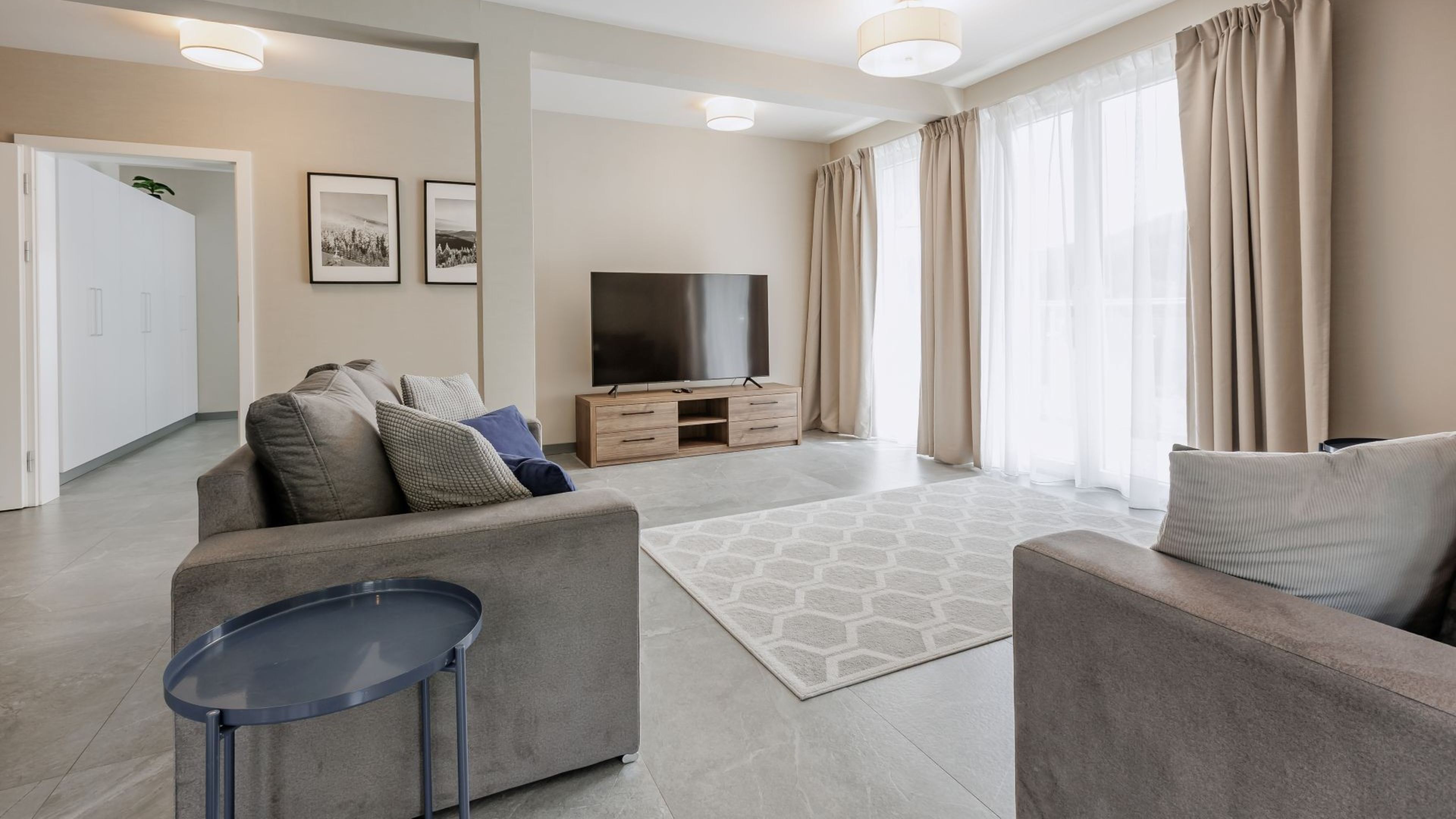Un salon moderne avec un canapé, une table basse et un téléviseur. Il y a des rideaux aux fenêtres et une porte qui donne sur une autre pièce.