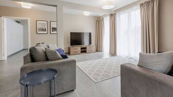 Un salon moderne avec un canapé, une table basse et un téléviseur. Il y a des rideaux aux fenêtres et une porte qui donne sur une autre pièce.