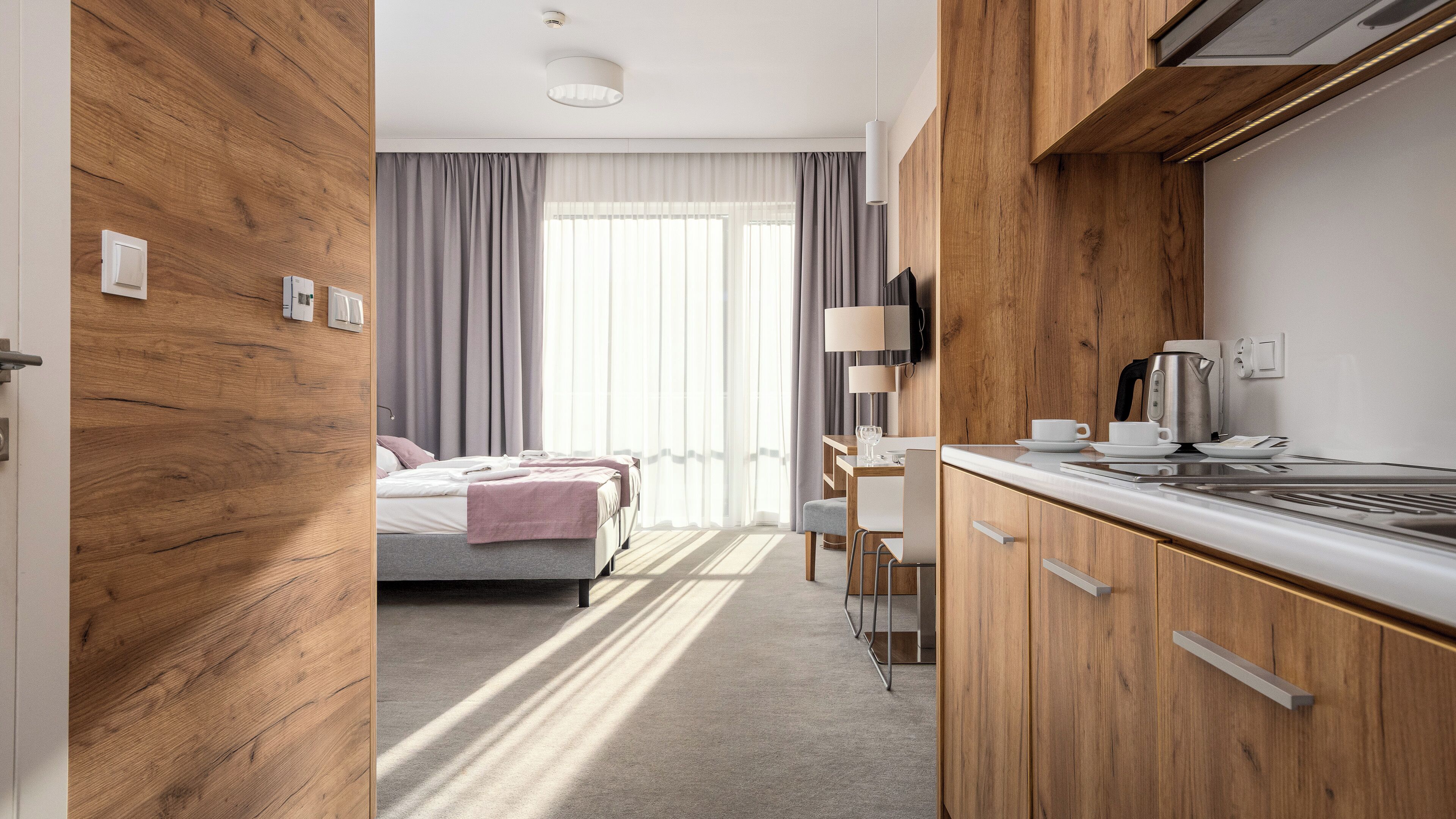 Una camera d'albergo moderna con una parete in legno decorativa, un letto con coperta rosa, una scrivania con lampada e un angolo cottura con macchina del caffè.