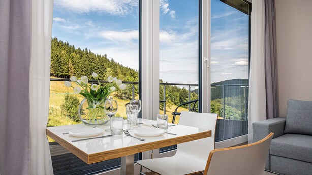 A dining area with a table set for two, a vase of white flowers, and a scenic view of a forested hillside through large windows. - Aparthotel Panorama – Czarna Góra Resort | Sun & Snow (Sienna)