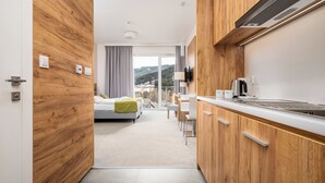 A modern hotel room with a wooden interior, a kitchenette, a bed with a green blanket, and a view of a forested landscape. - Aparthotel Panorama – Czarna Góra Resort | Sun & Snow (Sienna)