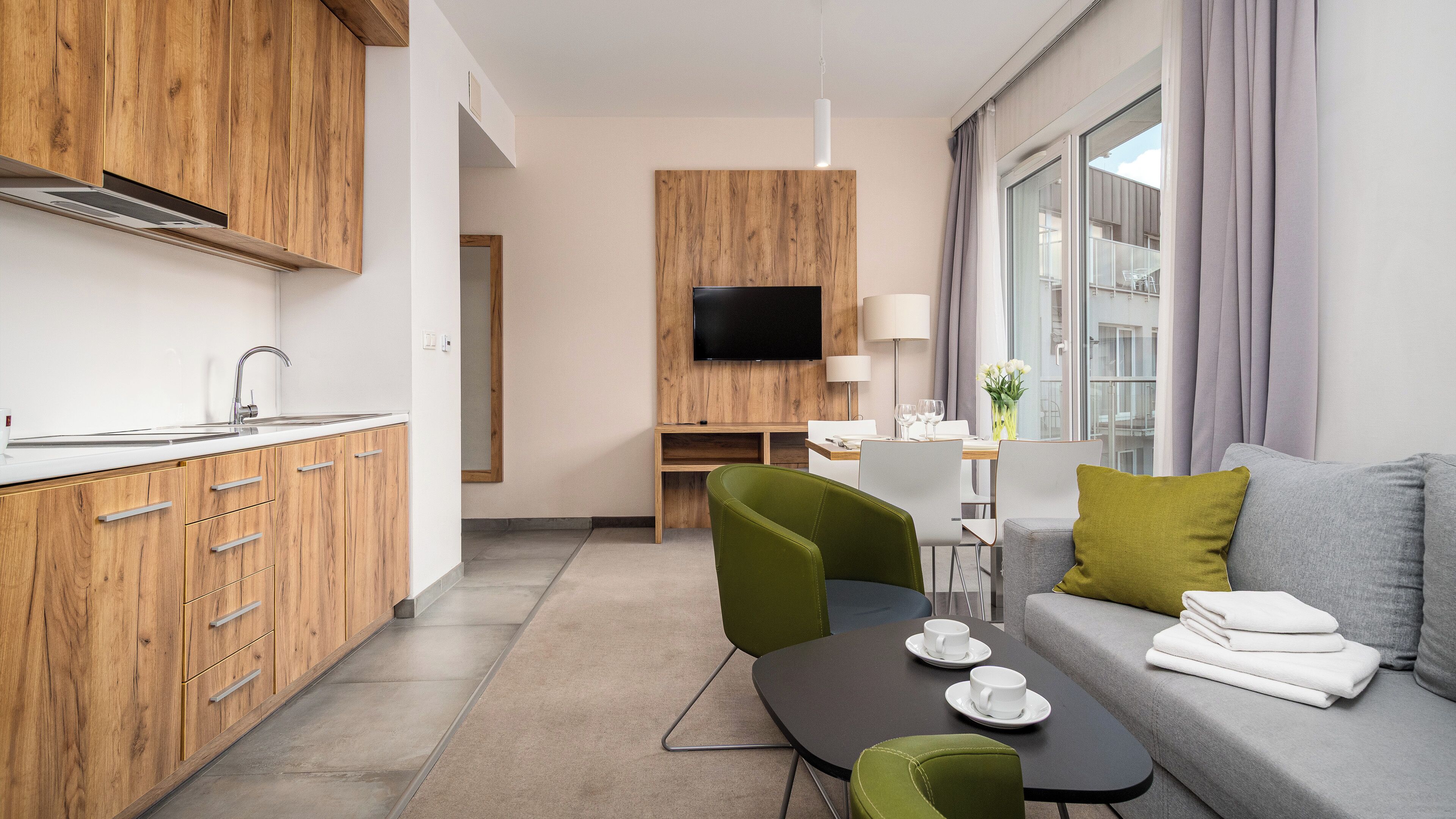 A modern hotel room with a kitchenette, a dining area, a sofa, and a TV.