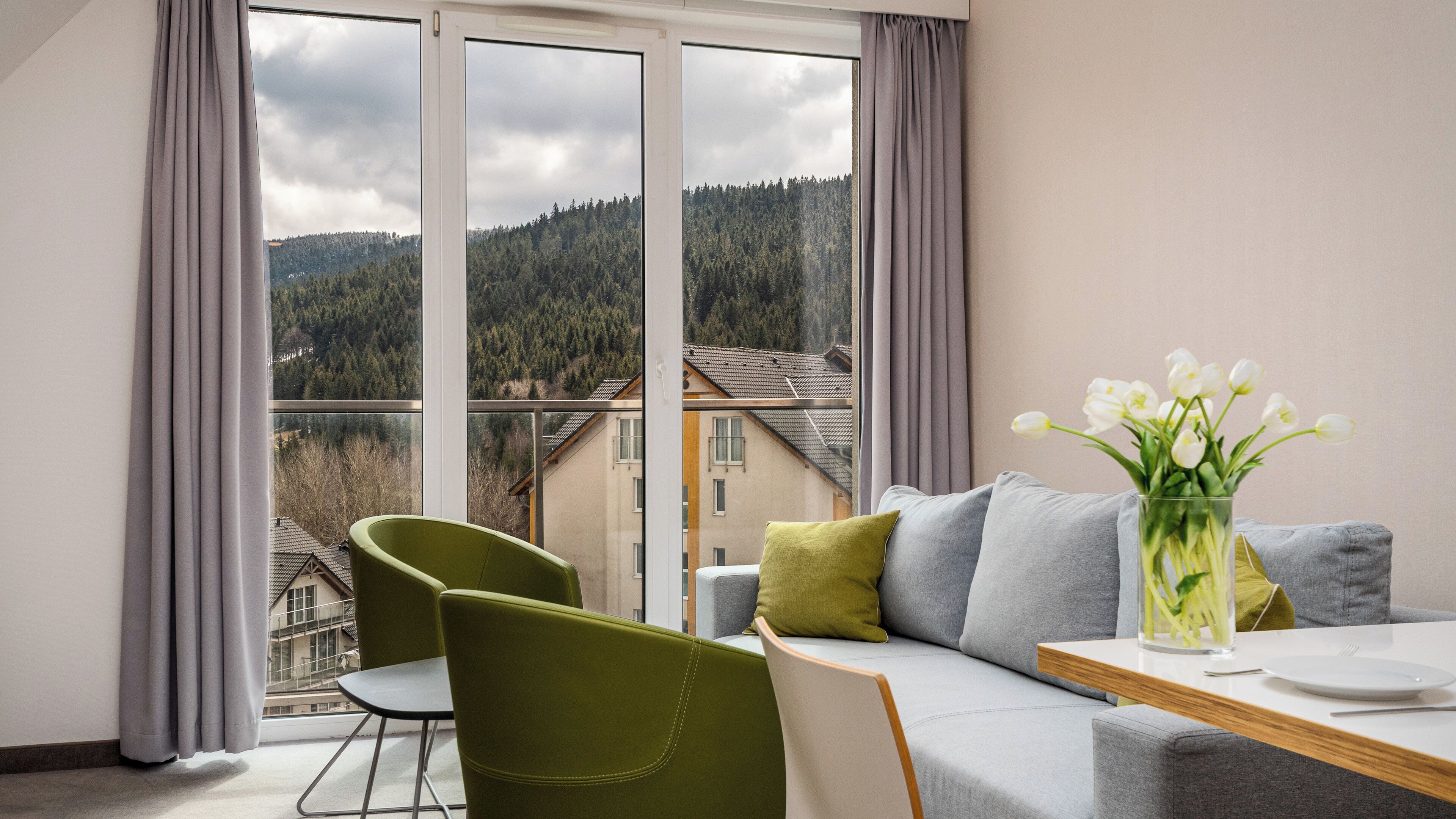 Una sala de estar moderna con sofá, mesa de comedor y sillas. Se ve un ventanal con vistas a una montaña boscosa y edificios.