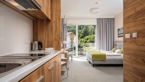 A modern hotel room with a kitchenette, a balcony with a view of greenery, and a bed with a green throw. - Aparthotel Panorama – Czarna Góra Resort | Sun & Snow (Sienna)