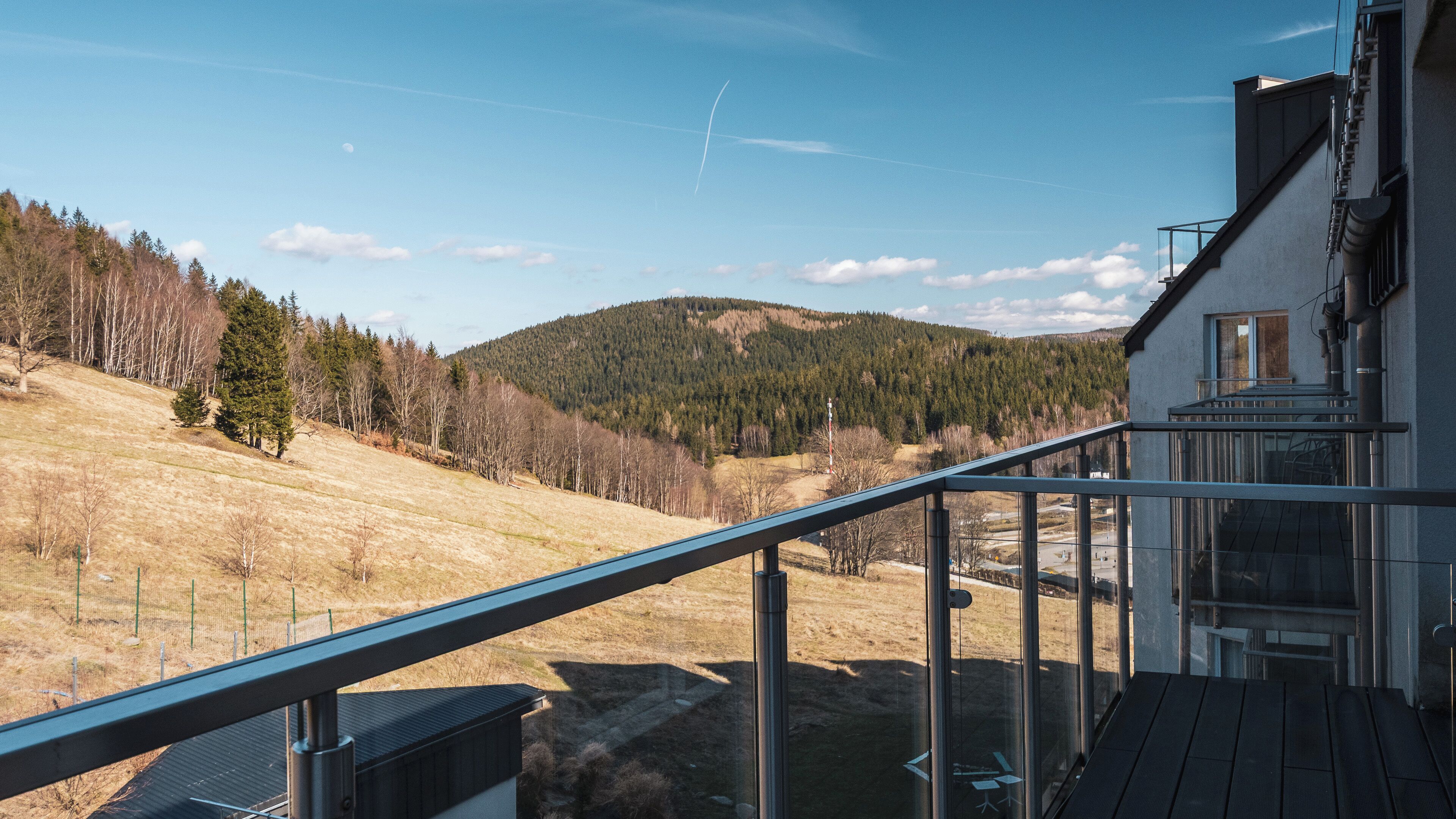 A balcony with a metal railing overlooking a hilly landscape with trees and a clear sky.