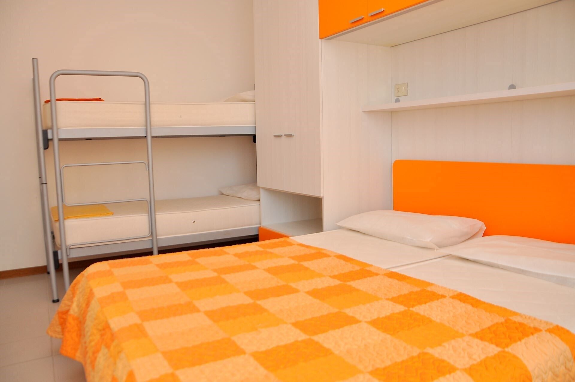 A bunk bed with a checkered orange and white blanket, a metal ladder, and a white wall with a shelf above.